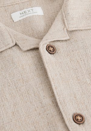 Camicia di lino beige con superficie testurizzata, caratterizzata da colletto, bottoni marroni e un'etichetta con la scritta "NEXT, MADE IN BANGLADESH."