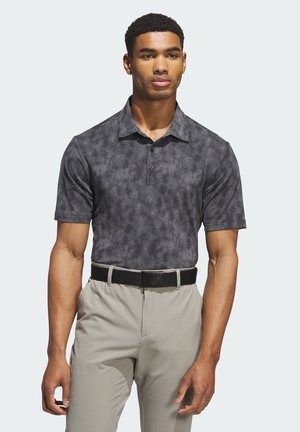 Muž stojící proti jednoduchému světlému pozadí, má na sobě tmavě šedé polo tričko s potiskem, béžové kalhoty a černý pásek.