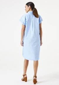 Hellblaues Baumwollkleid mit kurzen umgeschlagenen Ärmeln, einem Rückenschlitz und mittellangem Saum; kombiniert mit braunen Blockabsatzsandalen.