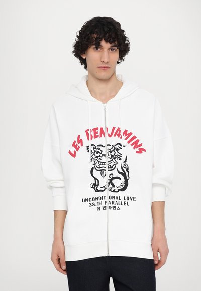 Λευκό φούτερ με κουκούλα και φερμουάρ, με γραφικό ενός λιονταριού και κείμενο σε κόκκινο και μαύρο. Φτιαγμένο από μαλακό, υφασματοποιημένο ύφασμα.