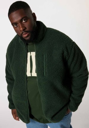 Green fleece jacket with a textured design, zip closure, and a chest pocket; worn over a green shirt with a light-colored graphic.