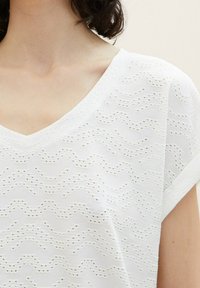 Žena v bílém topu z texturované látky s výstřihem do V a krátkým rukávem, který odhaluje krk a horní část hrudníku.