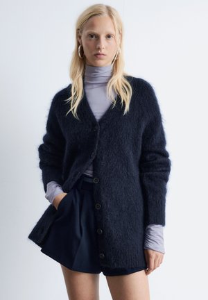 Marineblaue, flauschige Strickjacke mit V-Ausschnitt, zwei vorderen Taschen und schwarzen Knöpfen. Über einem helllila Langarmoberteil und Shorts getragen.