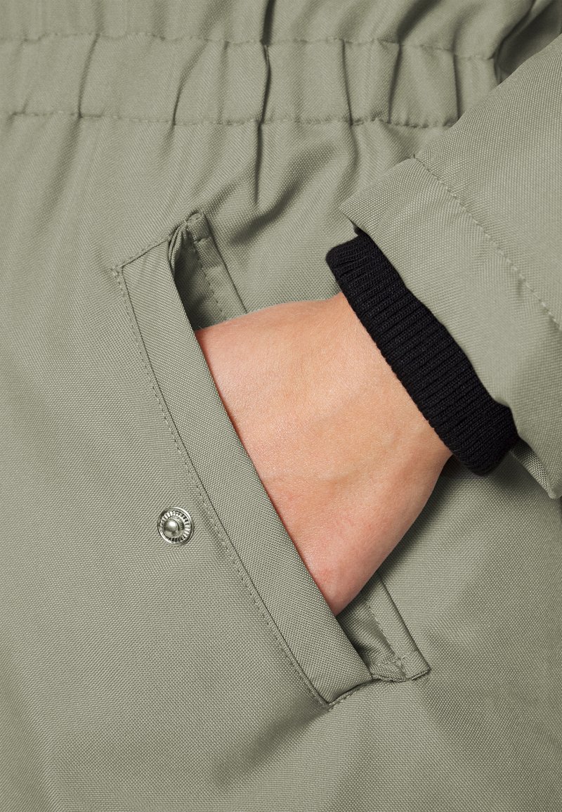 Poche de veste en tissu texturé vert avec une main à l'intérieur. Présente une fermeture par bouton-pression. Les poignets sont en tricot côtelé noir.