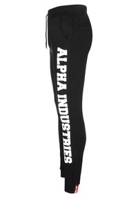 Zwarte joggers van zacht materiaal met witte "ALPHA INDUSTRIES" tekst op het linkerpijpje, voorzien van een contrasterend rood label aan de zoom en zakken.