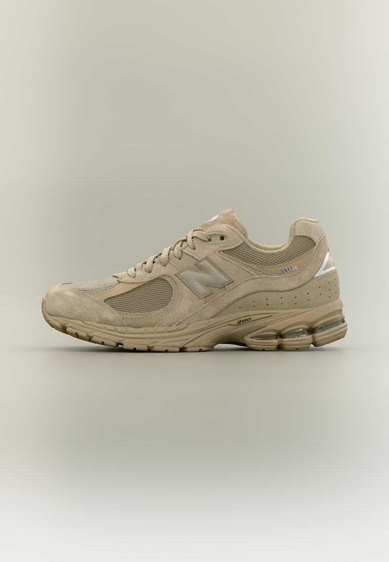 Μπεζ αθλητικό παπούτσι New Balance 2002R με επάνω μέρος από δίχτυ και σουέντ, απορροφητική σόλα και ανακλαστικές λεπτομέρειες σε ουδέτερο φόντο.