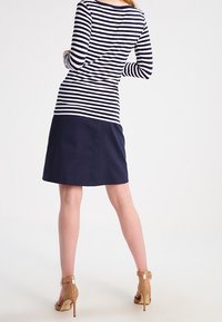 Haut à manches longues rayé avec un corps marine et une silhouette ajustée, associé à une jupe marine. Des chaussures à talons hauts beige avec des accents métalliques sont portées.