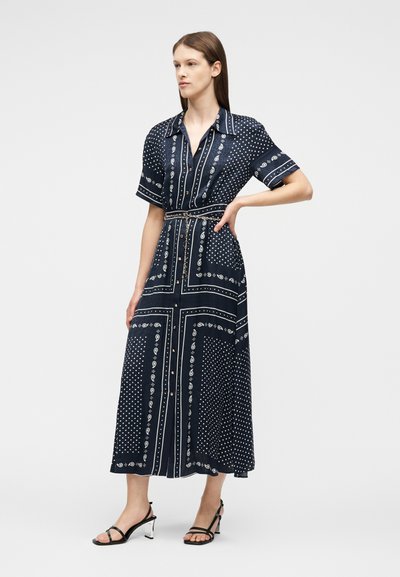 Γυναίκα που φοράει midi φόρεμα σε ναυτικό μπλε χρώμα με λευκά σχέδια πεϊсли και πουά, με κουμπιά μπροστά, κοντά μανίκια και μαύρα πέδιλα με λουράκια, όρθια με το χέρι στη μέση.
