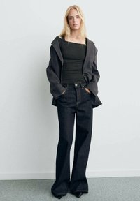 Débardeur noir à côtes sous un blazer gris surdimensionné, associé à un pantalon en denim foncé à pattes d'éléphant. Comprend des détails de poches et un matériel minimal.