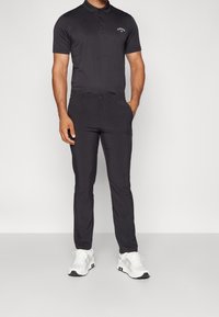 Černé polo tričko s límečkem a třemi knoflíky, v kombinaci s černými slim-fit kalhotami a bílými sportovními botami se šedými akcenty. Jednoduchý design.