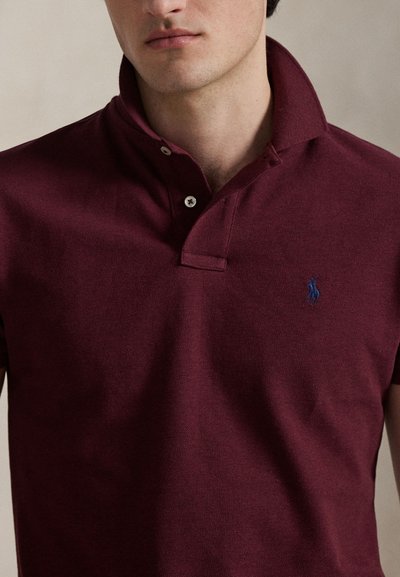 Homme portant un polo bordeaux avec un col relevé et un petit logo bleu brodé sur la poitrine gauche.