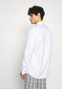 Camicia bianca a maniche lunghe con colletto classico, polsini con bottoni e tessuto morbido. Abbinata a pantaloni grigi a quadri.