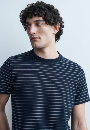 Νεαρός με καστανά σγουρά μαλλιά που φοράει μπλουζάκι με οριζόντιες μπλε και λευκές ρίγες, κοιτάζει προς τα αριστερά του.