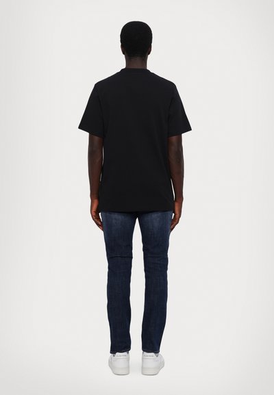 Sort t-shirt med korte ærmer, i bomuldsstof, afslappet pasform, parret med mørkeblå jeans og hvide sneakers, set fra bagsiden.