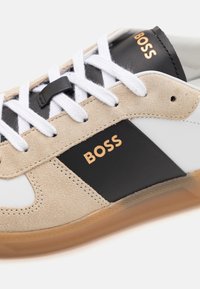 Sneaker con una combinazione di suede beige, pelle nera e pelle bianca. Presenta lacci bianchi e una suola in gomma trasparente. Logo "BOSS" in oro.