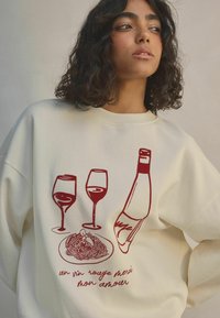 Sweatshirt crème avec un design graphique rouge représentant deux verres à vin, une bouteille de vin et une assiette de pâtes, accompagné de texte en dessous.
