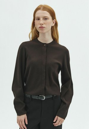 Giovane donna con lunghi capelli rossi che indossa un cardigan marrone scuro con bottoni e pantaloni neri con cintura nera, in piedi davanti a uno sfondo semplice.
