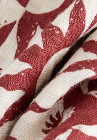 Gros plan d'un tissu texturé avec un motif floral rouge en relief sur fond crème, montrant le tissage et les plis détaillés.