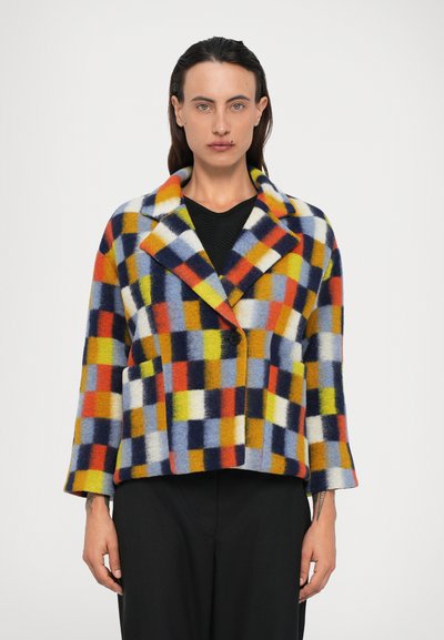 Femme portant une veste à carreaux multicolore avec un seul bouton noir sur un haut noir et un pantalon noir, se tenant devant un fond blanc.