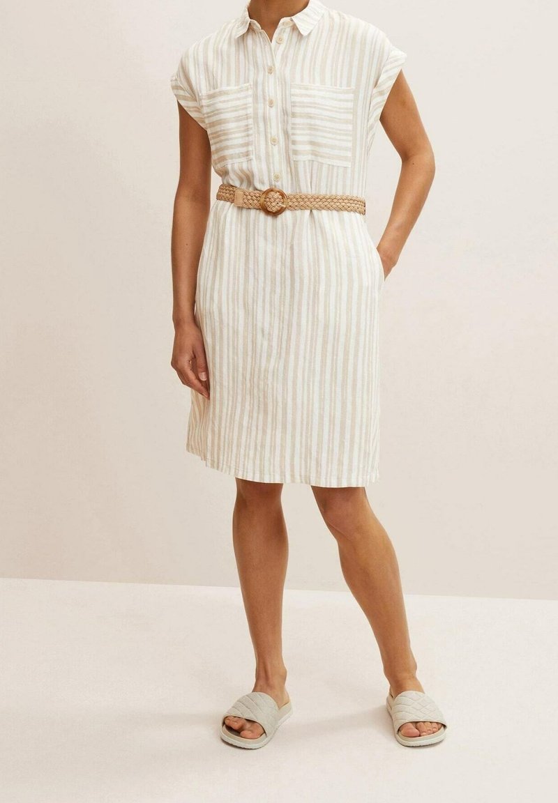 Femme portant une robe beige et blanche à rayures verticales jusqu'aux genoux, avec des manches courtes, des poches avant, une ceinture tissée et des sandales matelassées.