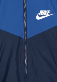 Nike větrovka s modrou horní částí a tmavě námořnickou spodní částí. Obsahuje centrální zip a bílé logo v levém horním rohu.