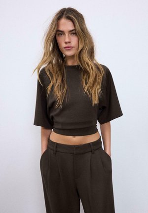 Femme aux longs cheveux ondulés portant un haut court sombre et un pantalon taille haute assorti, debout les mains dans les poches devant un fond clair.