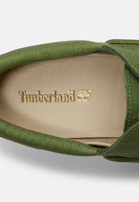 Chaussure en cuir verte avec col rembourré et logo Timberland doré sur une semelle intérieure beige, mettant en avant la marque et les détails de la texture.
