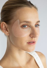 Gennemsigtige underøjen-patches på en models ansigt, der fremhæver en glat hudstruktur og naturlig makeup. Lys baggrund.