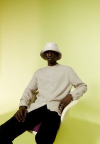 Beige overhemd met lange mouwen en een mandarijnkraag, gedragen met een lichtgrijze bucket hat met een logo. Zwarte broek, zittend op een stoel.