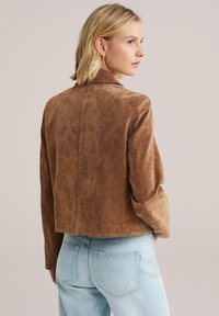 Bruine suède jas met een gestructureerde afwerking, voorzien van een kraag en een cropped ontwerp. Gecombineerd met lichtblauwe spijkerbroek.