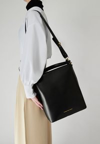 Svart läderväska med strukturerad design, guldfärgade detaljer och en lång justerbar rem, hålls av en person i en vit blus.