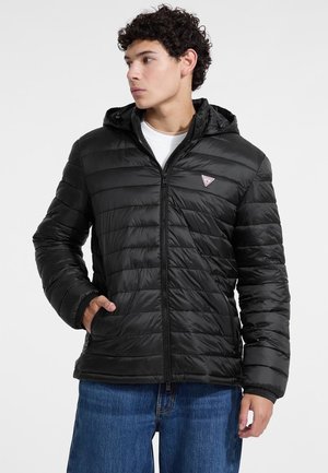 Black puffer jacket with hood, quilted texture, front zipper, side pockets, and small logo patch on left chest. Worn over a white shirt.