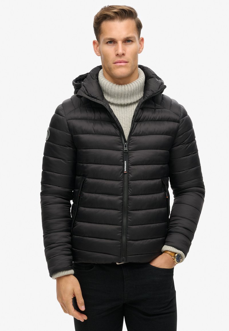 Chaqueta acolchada negra con cierre de cremallera y capucha. Presenta un cuello alto de canalé gris, bolsillos laterales y un acabado texturizado.