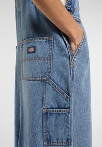 Džínové lacláče v světlé modré barvě s boční kapsou a značkovou nášivkou. Materiál vykazuje opotřebovaný vzhled s viditelným šitím.