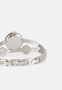 Sidabrinis metalinis laikrodis su apvaliu ciferblatu ir grandinėle. Yra išgraviruoti "GUESS" logotipai ir auksinė akcento detalė ant karūnėlės. Lygi textura.