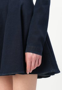 Donkerdenim jurk met een aansluitend lijfje en een uitlopende rok. Close-up toont details van een omgeslagen mouw en een rauwe zoom.