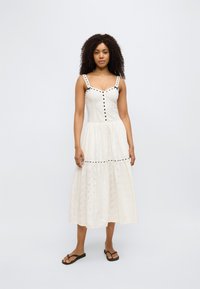 Λευκό βαμβακερό φόρεμα με διάτρητο σχέδιο, εφαρμοστό μπούστο και φούστα με στρώσεις. Μαύρα κουμπιά κατά μήκος του μπροστινού μέρους και τιράντες με αντίθετες μαύρες λεπτομέρειες.