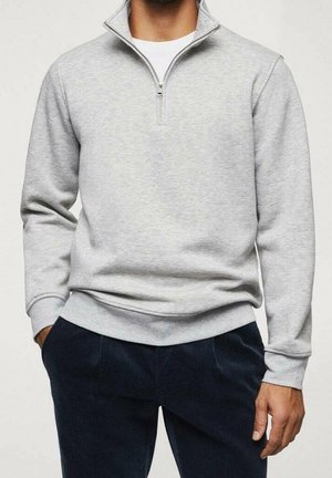 Hombre con sudadera de media cremallera de color gris claro sobre camisa blanca y pantalones oscuros, de pie con las manos en los bolsillos.