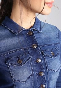 Jeansjacka med en mörk tvätt, med en klassisk krage, metallknappar och två bröstfickor. Sömmar ger detaljer till designen.