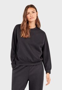 Sudadera negra con cuello redondo, fabricada en tejido suave, con puños y dobladillo acanalados, corte relajado y bolsillos laterales. Combinada con pantalones negros a juego.