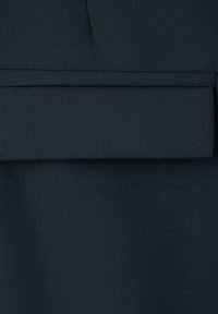 Gros plan d’un vêtement en tissu foncé avec un rabat horizontal en tissu et un détail de couture au-dessus.