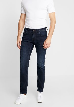 Mężczyzna ubrany w ciemnoniebieskie dopasowane jeansy, białą koszulkę i białe trampki stoi na jasnym jednolitym tle.