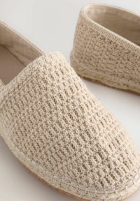 Zapatos slip-on beige de tejido trenzado con un upper de tela texturizada, presentan una suela plana y detalles de costura en el borde.