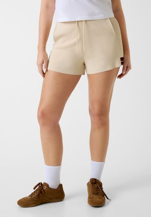 Femme portant un short en tricot beige avec cordon de serrage, des chaussettes blanches, des baskets marron, et un haut court blanc, debout sur un fond uni.