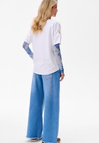 Biała bluzka z krótkim rękawem, z niebieskimi długimi rękawami tie-dye, w połączeniu z szerokimi, jasnoniebieskimi dżinsami z frędzlami na końcach.