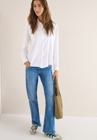 Femme portant une chemise blanche boutonnée, un jean bleu et des baskets, tenant un sac cabas vert matelassé à côté d'elle, debout devant un fond uni.