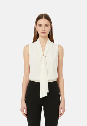 Donna con capelli lisci che indossa una camicetta bianca senza maniche con cravatta e pantaloni neri, in piedi davanti a uno sfondo chiaro e uniforme.