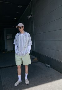 Chemise à manches courtes rayée avec fermeture à boutons, shorts à carreaux verts clairs, chaussettes blanches et chaussures tissées beiges, sur fond en béton.