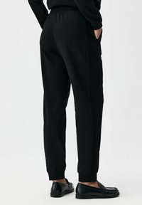 Czarne spodnie dresowe z miękkiego materiału, wyposażone w boczne kieszenie, ściągacze przy mankietach oraz elastyczny pas. Noszone z czarnymi mokasynami.