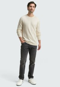 Mann steht entspannt mit lockeren Händen, trägt einen beigen Pullover, dunkelgraue Jeans und weiße Turnschuhe vor einem schlichten hellen Hintergrund.